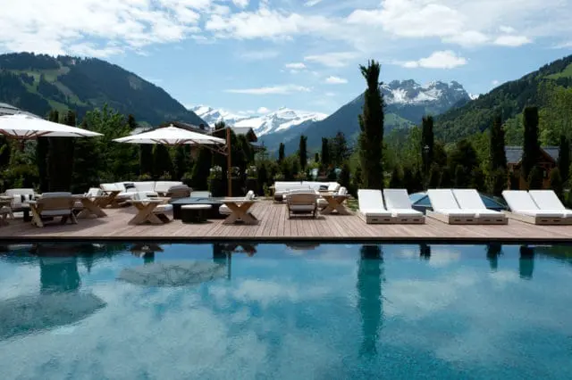 Outdoor swimming pool with lounge chairs and seating area on wooden deck, surrounded by trees and mountains under a cloudy sky