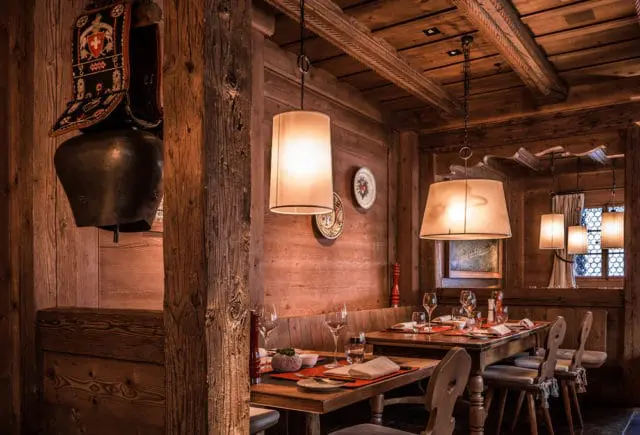 Rustic wooden restaurant interior with tables set for dining, featuring a large cowbell and Swiss flag decoration on the wall, and warm pendant lighting.