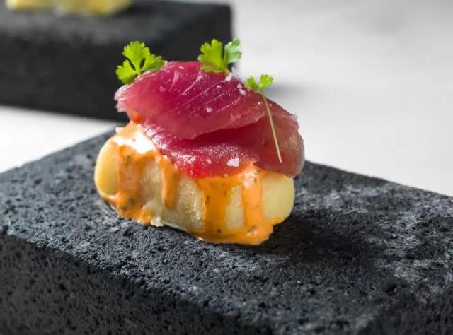 Tuna causa appetizer with raw tuna slices, creamy orange sauce, and parsley garnish served on a dark, textured stone slab.