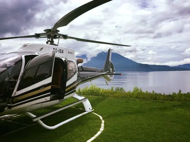 White helicopter with black and gold stripes parked on a grassy helipad next to a large lake and mountains under a cloudy sky.