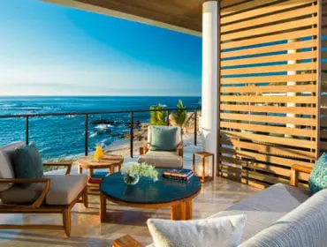 Outdoor living room with wooden furniture and slatted wood privacy screen overlooking the ocean and rocky coastline.