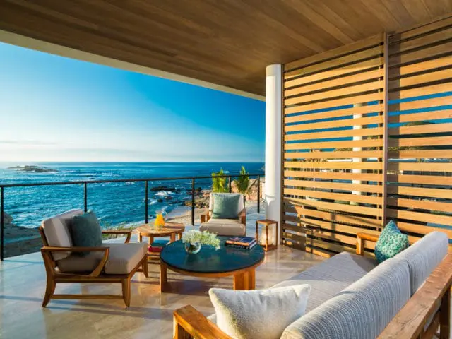 Covered outdoor living room with wooden slatted walls, comfortable seating, and a view of the ocean and rocky coastline.