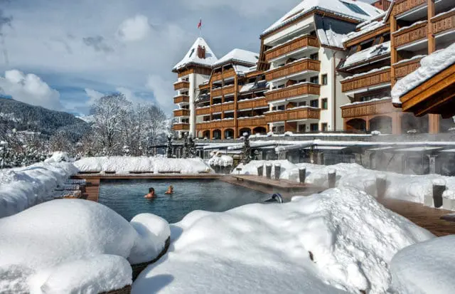 Luxury hotel with wooden balconies and white facade covered in snow, overlooking a steaming outdoor pool with two people swimming in winter.