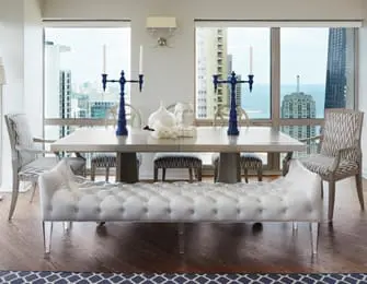 Dining room with a tufted white bench, a light wood table, and upholstered chairs, set against a backdrop of city skyline views through large windows.