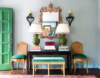 Entryway with a bright green door, a black console table topped with two green pineapple lamps and books, flanked by two gold chairs with teal velvet seats, and a decorative mirror above.