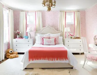 Bedroom with pink and white spotted wallpaper, a white upholstered headboard bed with coral throw blanket, two white nightstands, and a gold chandelier.