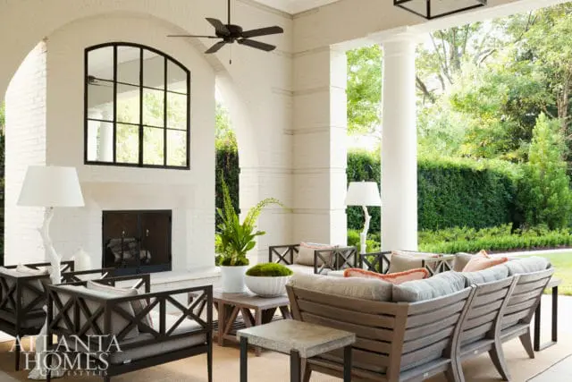 Covered porch with white brick fireplace, arched window, ceiling fan, and outdoor seating area with wooden furniture and plush cushions.