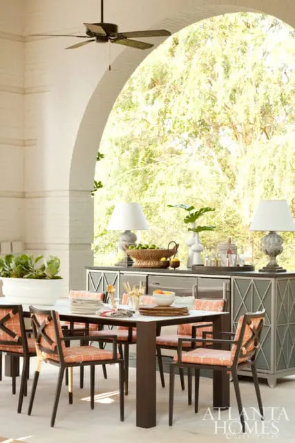Covered patio dining area with a white table set for meals, surrounded by chairs with orange and white patterned cushions. A dark gray cabinet with diamond pattern doors and built-in appliances is visible behind the table.