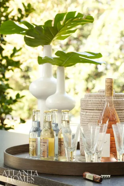 White vases with monstera leaves, bottles of tonic water and rose wine, and glasses on a tray with a woven basket in the background.