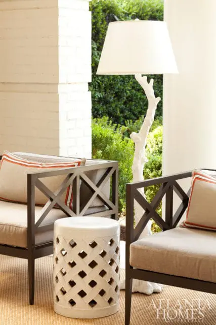 Outdoor seating area with dark metal chairs and beige cushions, a white lattice garden stool, and a white tree-trunk lamp with a cream shade.