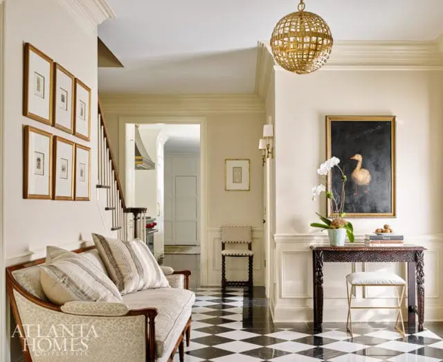 Hallway with black and white checkered floor, a patterned settee, framed artwork on the wall, and a painting of a duck above a carved console table.