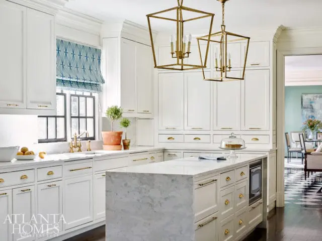 White kitchen with marble island, brass hardware, and geometric blue roman shades over a window. Two gold lantern pendant lights hang above.