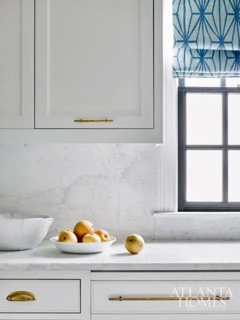 White kitchen with marble countertops and backsplash, gold hardware on cabinets and drawers, and a blue geometric patterned window shade.