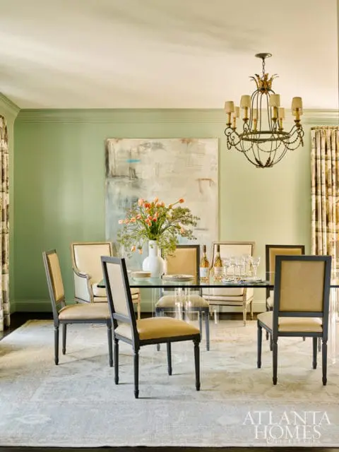 Dining room with pale green walls, glass dining table, upholstered chairs with dark wood frames, abstract art, and ornate chandelier.