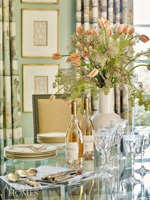 Glass dining table set with stacked gold-rimmed plates, silverware, two bottles of rosé wine, crystal wine glasses, and a vase of peach tulips and greenery.