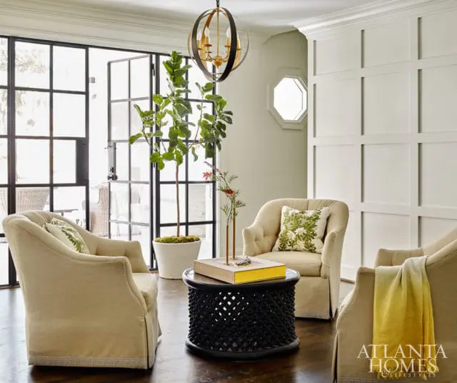 Living room with three upholstered armchairs, a dark wood carved coffee table with a yellow book, and a large potted fiddle leaf fig tree.