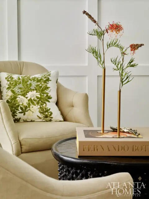 Tufted cream armchair with green floral pillow next to a dark carved wood table holding a book and brass vases with dried flowers.