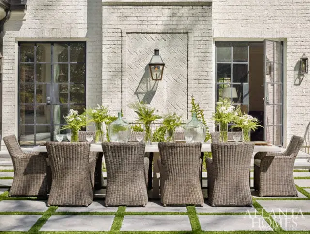 Wicker dining chairs surround a long table on a patio with white brick walls, large metal-framed windows, and glass doors. Greenery and glass vases adorn the table.