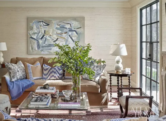 Living room with a tan sofa, abstract art, patterned pillows, and a glass coffee table filled with books. A large window with black mullions is visible on the right.