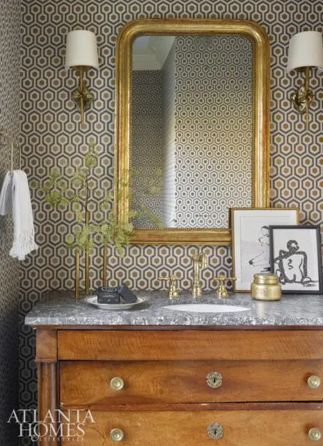 Powder room with hexagonal patterned wallpaper, marble countertop, antique wooden vanity, gold-framed mirror, brass fixtures, and decorative artwork.