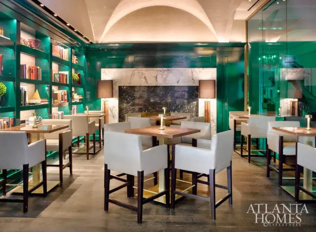 Bar area with white leather stools and wood-topped tables, flanked by emerald green lacquered walls with built-in bookshelves and a marble fireplace.