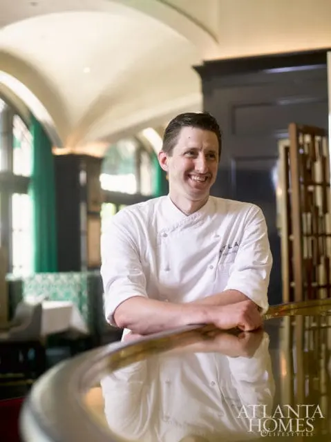 Chef in white uniform smiling with arms crossed, leaning on a polished bar in a restaurant with green accents and arched windows.