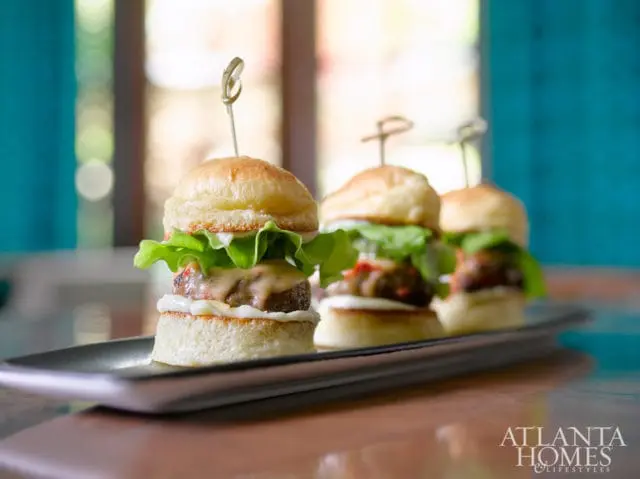 Three gourmet sliders on a dark rectangular platter, topped with lettuce, cheese, and a skewer.