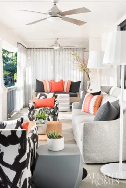 Covered porch with white ceiling, two ceiling fans, and sheer curtains. Furniture includes a sofa, patterned armchairs, and accent pillows in orange and pink.