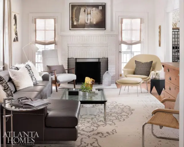 Living room with a dark leather sofa, white armchair, cream-colored lounge chair with ottoman, glass coffee table, and a white brick fireplace.