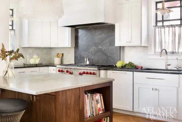 White kitchen with a marble island, stainless steel range with red knobs, dark stone backsplash, and white cabinetry with diamond-patterned tile backsplash.