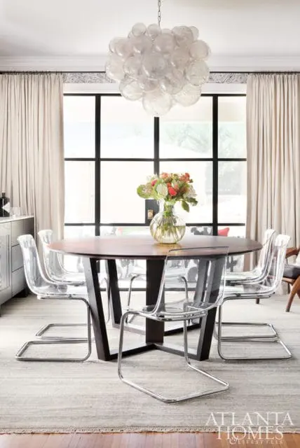 Round wood dining table with clear acrylic chairs and a bubble chandelier, set against a large window with beige curtains.