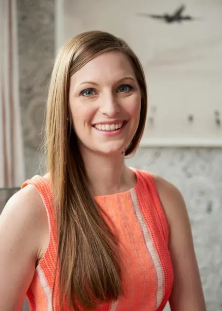 Woman with long brown hair and blue eyes smiling, wearing an orange sleeveless dress.