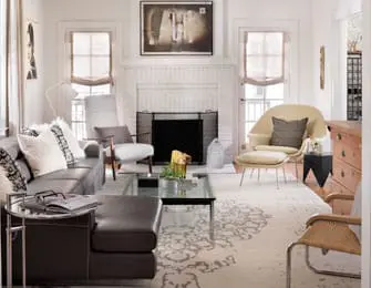 Living room with a dark gray sectional sofa, cream armchair with ottoman, and a glass-top coffee table on a patterned rug. A white brick fireplace is centered on the wall.
