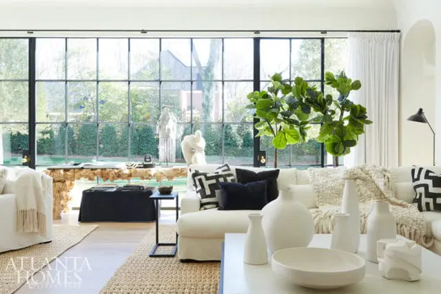 Living room with white sectional sofa, black and white patterned pillows, and a large fiddle leaf fig plant. Floor-to-ceiling windows overlook a garden.
