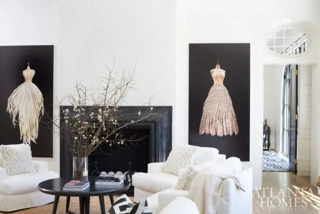 Living room with white sofas, black coffee table, and two large artworks of mannequins in dresses on black backgrounds. A fireplace and doorway are visible.