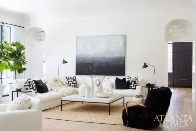 White living room with a large abstract painting, white sofas with black and white patterned pillows, a white coffee table with vases, and a black furry armchair.