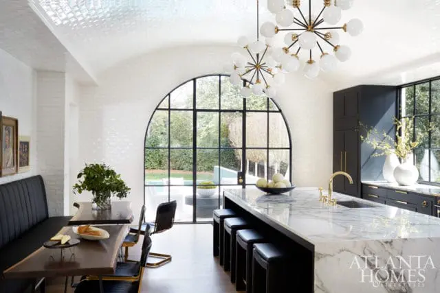 Modern kitchen with white subway tile walls, a large arched window overlooking a pool, marble island with black stools, and a dark wood dining table with a black banquette.