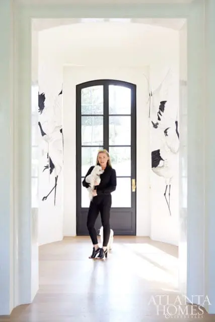 Woman holding a white dog in a hallway with a black arched door and large crane-themed wallpaper panels.