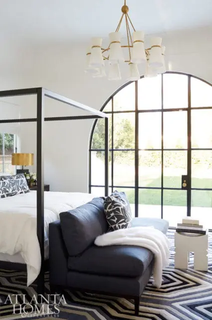 Bedroom with a black canopy bed, dark blue chaise lounge, and a large arched window overlooking a lawn. A gold chandelier hangs from the ceiling.