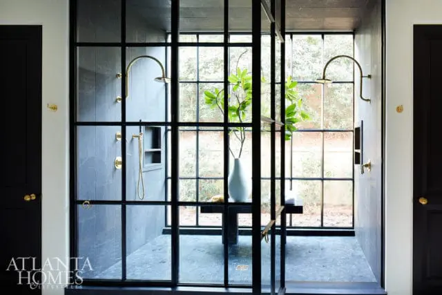 Modern bathroom with black metal-framed glass shower enclosure, gray stone tile walls, gold fixtures, and a large window overlooking greenery.