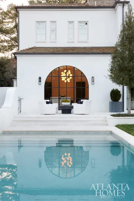 White stucco house with arched window and pool, featuring a patio with white chairs and a dark coffee table.