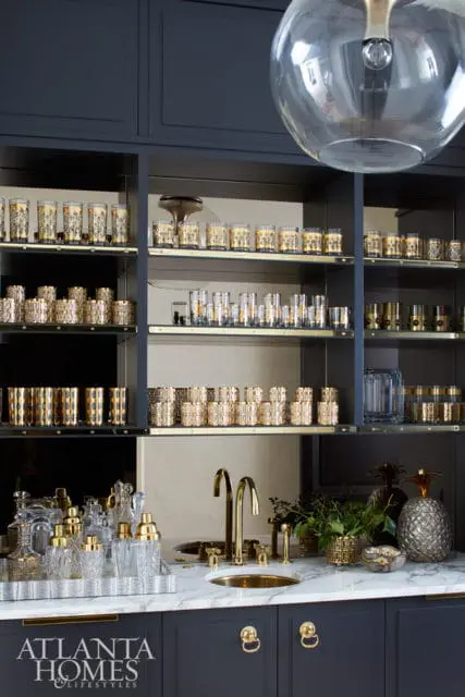 Bar area with dark blue cabinetry, marble countertop, gold faucet, and shelves filled with glassware and barware.