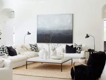 Living room with white sectional sofa, black and white patterned pillows, textured throw blanket, large abstract art, and a white stone coffee table with vases.