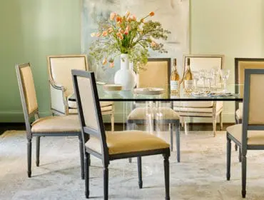 Dining room with a glass-top table, upholstered chairs in tan and cream, and a large abstract painting on a pale green wall.