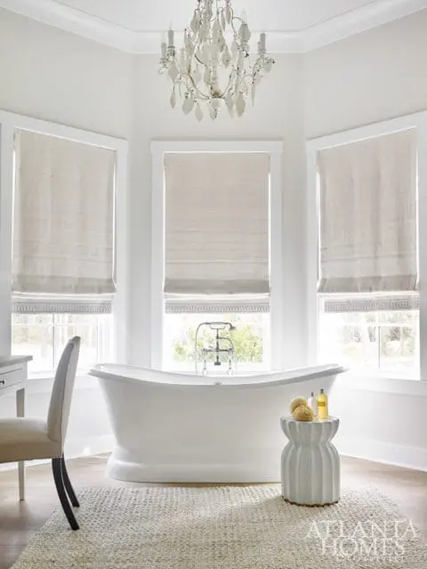 Bright white bathroom with a freestanding soaking tub, a decorative chandelier, and three windows with linen Roman shades.