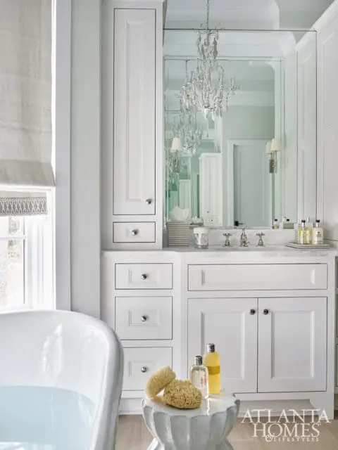 White bathroom with a freestanding tub, marble vanity with white cabinetry, and a crystal chandelier reflected in the mirror.