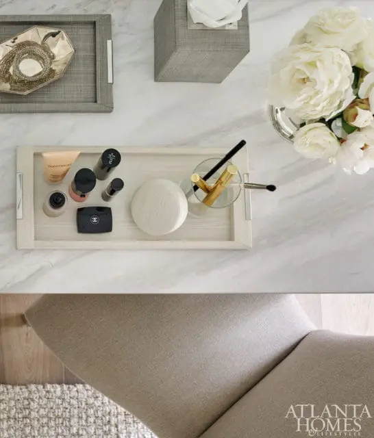 Top-down view of a vanity with a marble countertop, featuring a gray woven tray with jewelry, a gray tissue box, a tray with makeup and brushes, and a vase of white peonies.