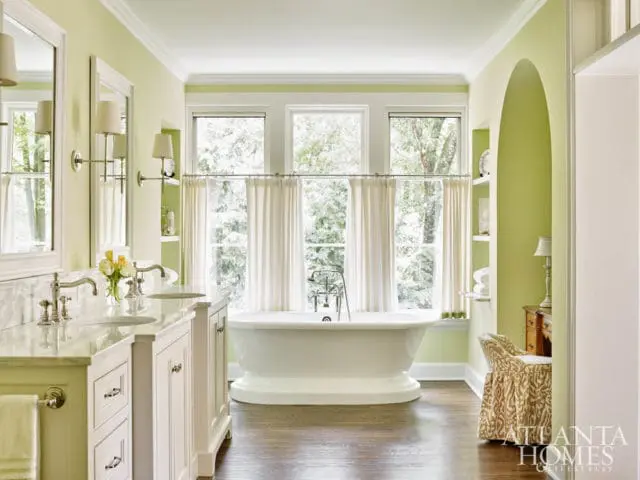 Light green bathroom with white double vanity, marble countertops, freestanding bathtub, and a patterned upholstered chair.