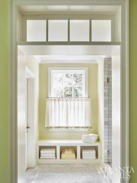 Light green bathroom with a window, white curtains, built-in shelves with folded towels, and marble tile floor.