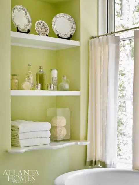 Pale green bathroom with white shelves displaying decorative plates, bottles, jars, and stacked towels. A glass vase holds two natural sponges. White curtains hang beside a window.